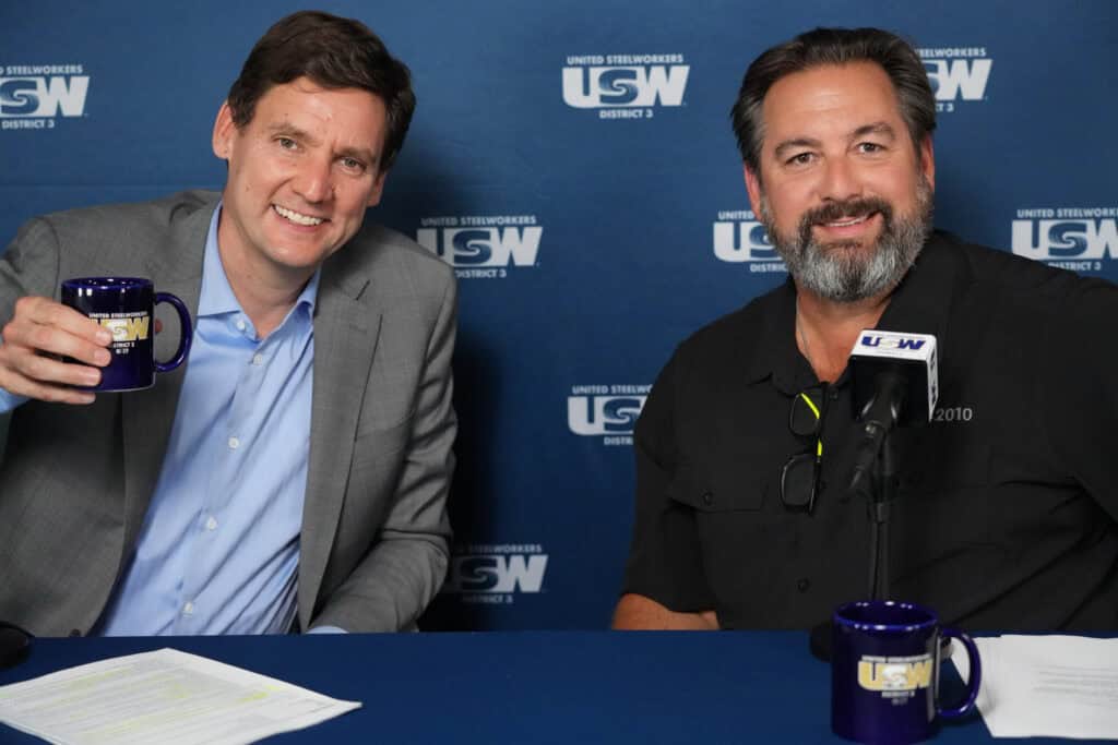 Two people are seated at a table facing the camera, smiling. The one on the left is wearing a suit jacket and dress shirt (no tie) and raising a coffee mug. Behind them is a dark blue backdrop with copies of the USW logo in white.