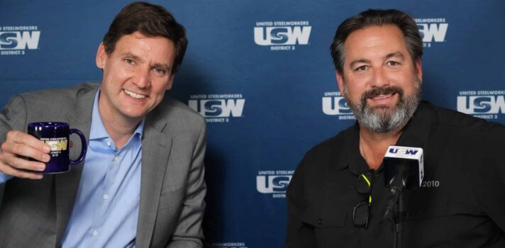 Two people are seated at a table facing the camera, smiling. The one on the left is wearing a suit jacket and dress shirt (no tie) and raising a coffee mug. Behind them is a dark blue backdrop with copies of the USW logo in white.