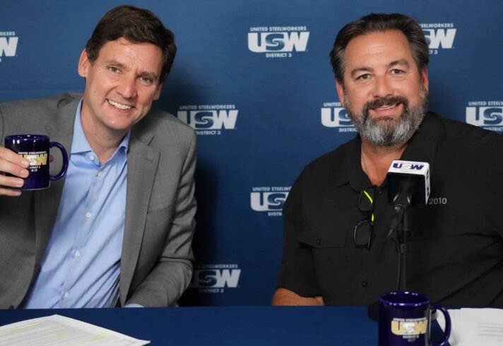 Two people are seated at a table facing the camera, smiling. The one on the left is wearing a suit jacket and dress shirt (no tie) and raising a coffee mug. Behind them is a dark blue backdrop with copies of the USW logo in white.