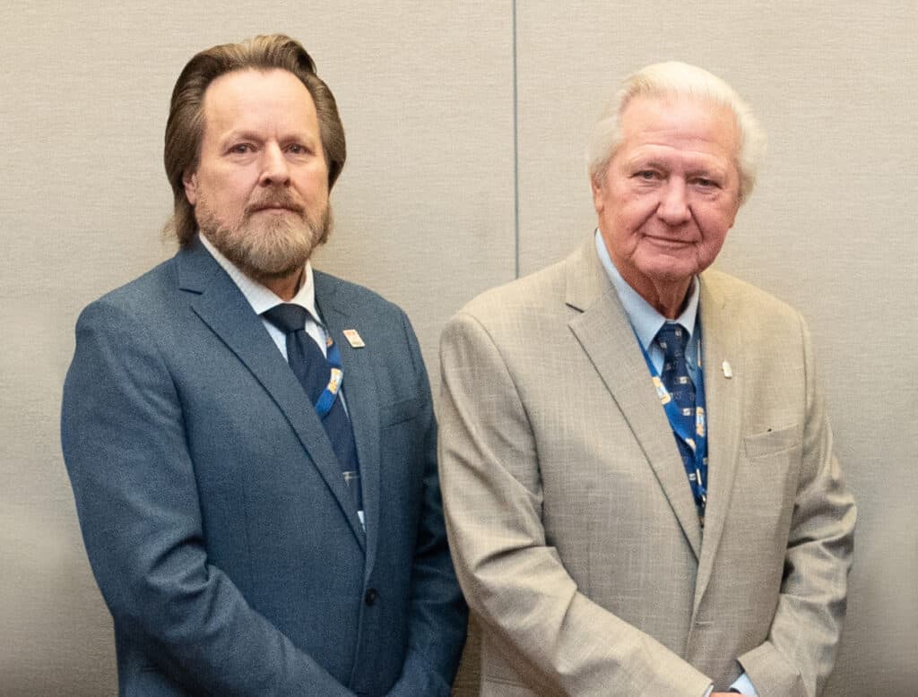 Two people in suit jackets stand side by side facing the camera with their hands folded in front of them.