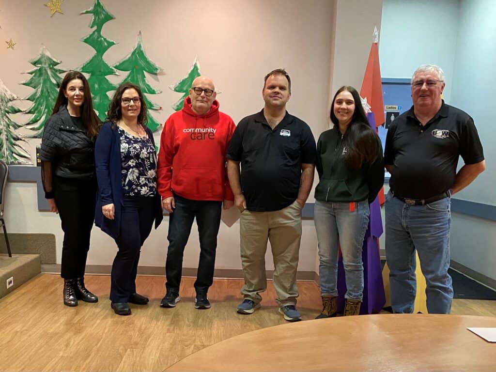 Six people stand beside each other indoors in a hall decorated for the holidays, facing the camera. One person's sweatshirt says "Community care"; another's has a USW logo on the shirt.