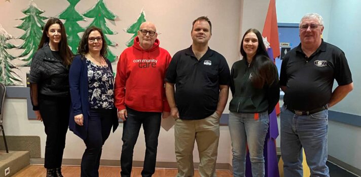 Six people stand beside each other indoors in a hall decorated for the holidays, facing the camera. One person's sweatshirt says 