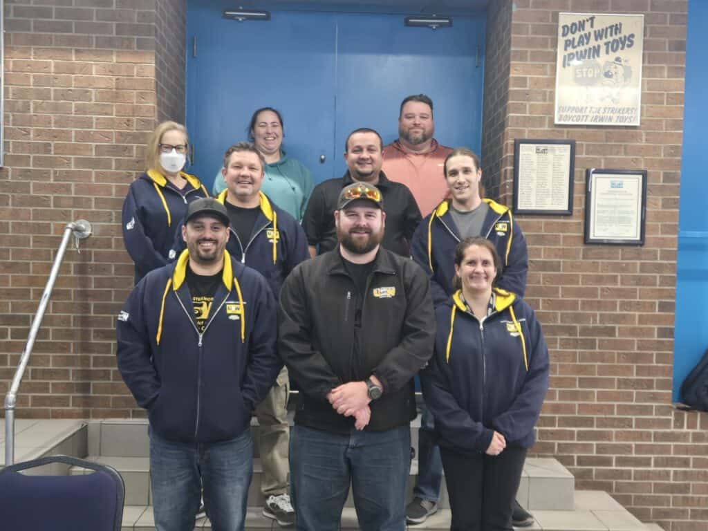 Nine people are standing and smiling at the camera in a classroom, with a blue door in the background and a brick wall behind them