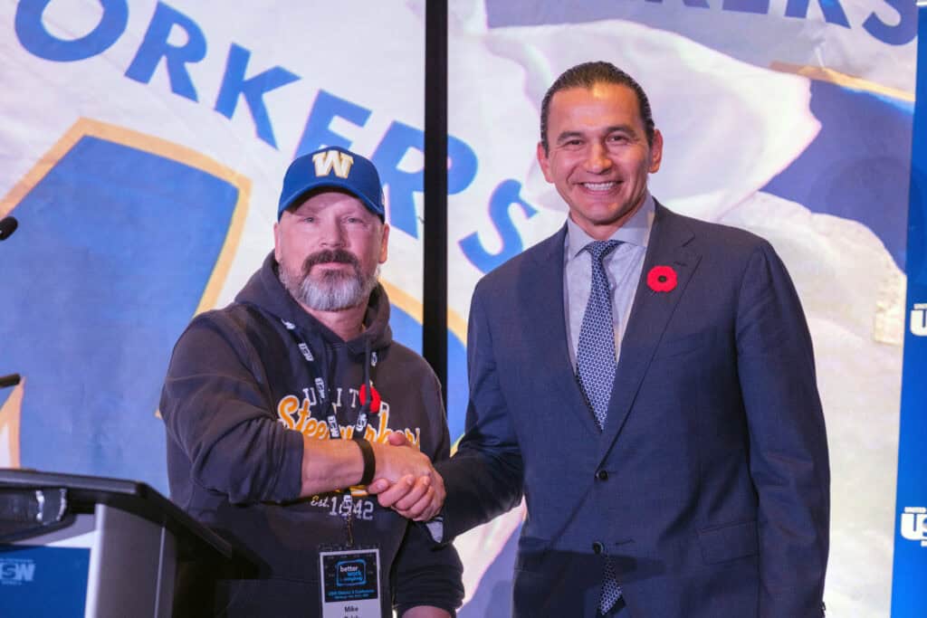 Mike Pulak shaking hands with Premier Wab Kinew in front of USW flag background