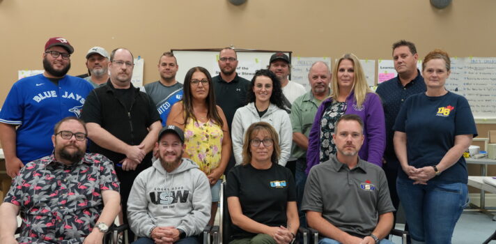 Sixteen people are standing in a classroom setting.