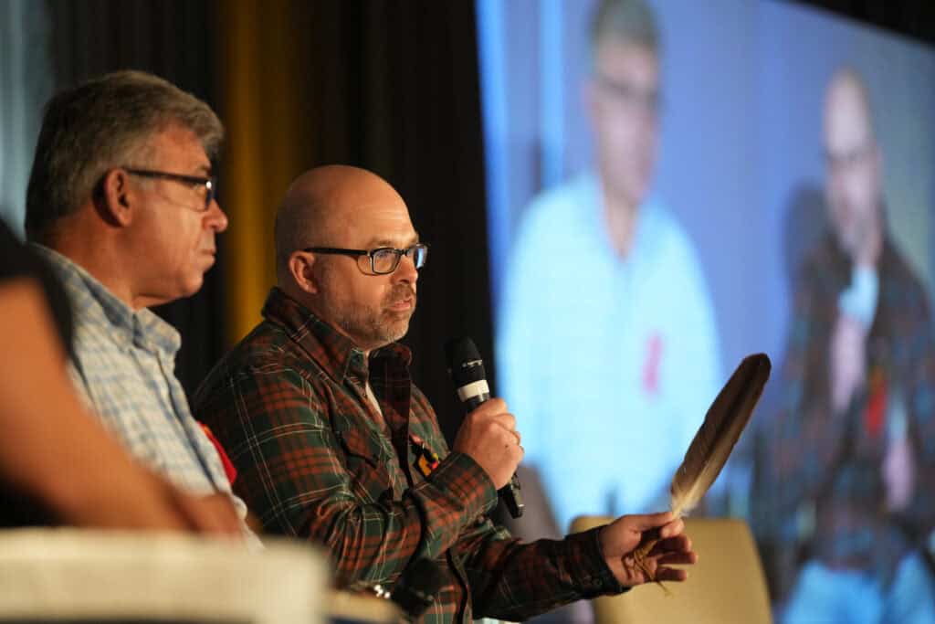 Two people both wearing glasses. One person is holding a feather and a mic while speaking to the crowd.