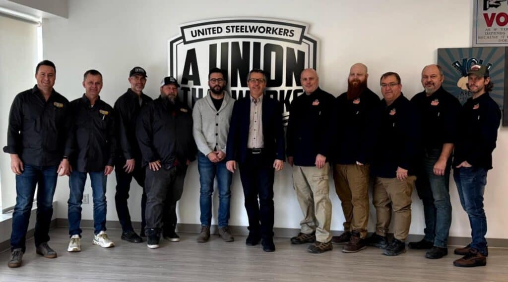 11 people standing in a room behind a United Steelworkers logo.