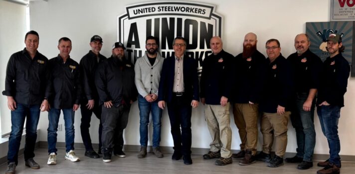 11 people standing in a room behind a United Steelworkers logo.