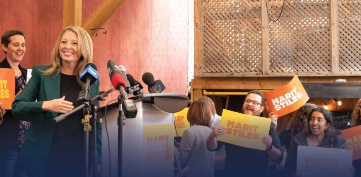 A photo of a person standing on stage during a press conference while smiling to the audience. There are people behind the stage holding signs with text saying Marit Stiles. There is a bold text under the photo saying: USW is proud to endorse MArit Stiles and Ontario NDP.