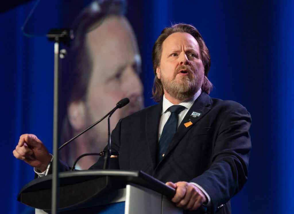 Image: man stands at microphone while speaking at a podium. They are dressed in a suit.