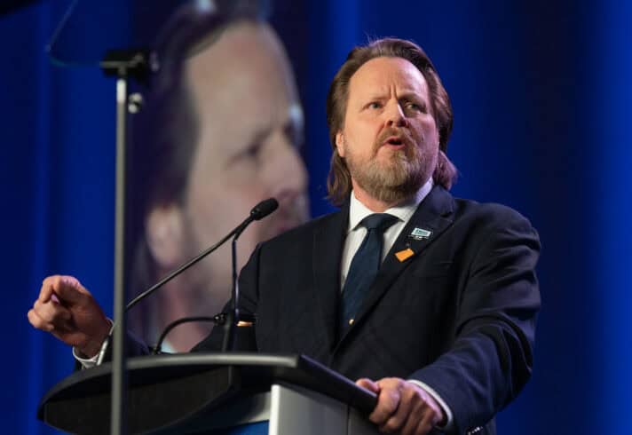 Image: man stands at microphone while speaking at a podium. They are dressed in a suit.