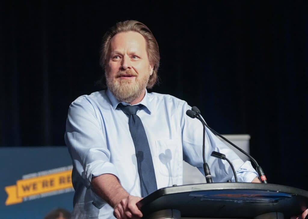 Image: man stands at microphone in front of large conference of 500 people.