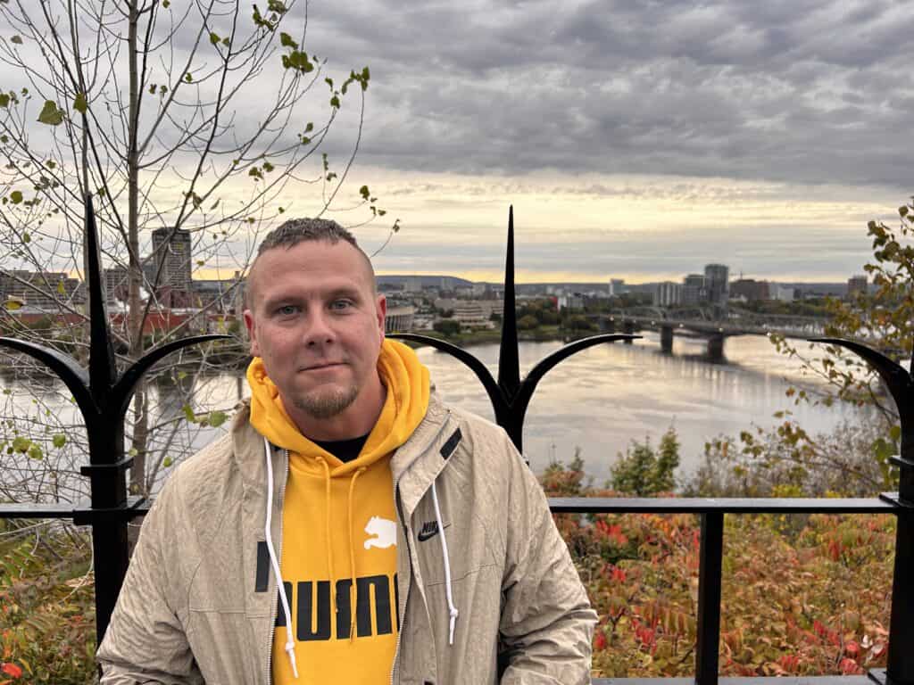 A man in a yellow sweater and beige jacket is standing outside and smiling.