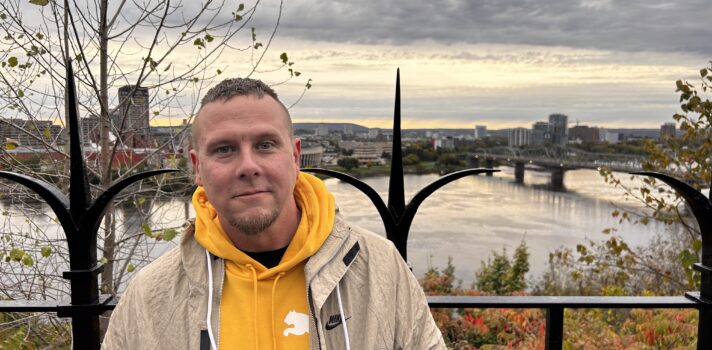 A man in a yellow sweater and beige jacket is standing outside and smiling.