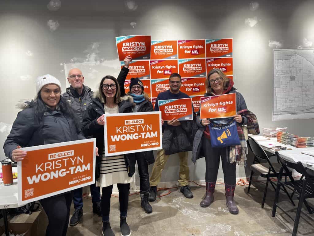 A group of people in a campaign office holding orange campaign signs.