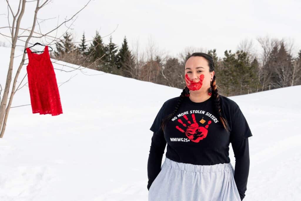 A person standing behind snow with a red dress hanging on a tree and red hand print on her face.