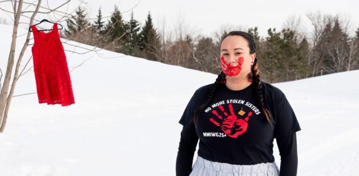 A person standing behind snow with a red dress hanging on a tree and red hand print on her face.