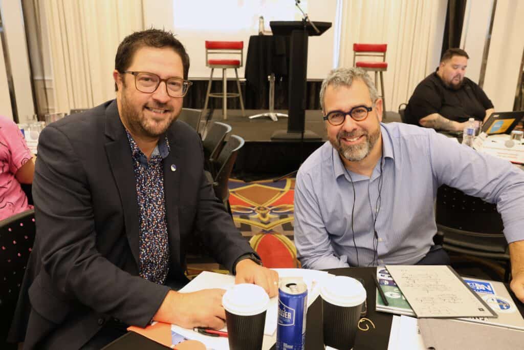 A man wearing glasses and a black vest sits at a table stacked with papers and coffee, smiling at the camera. Next to him sits another man wearing glasses and a light blue dress shirt; he's smiling.