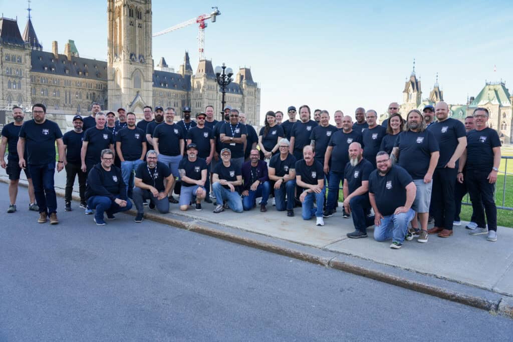 A group of over 30 people are gathered outdoors on Parliament Hill all wearing navy blue t-shirts. Some are standing, some are kneeling in front so those behind can be viewed by the camera.