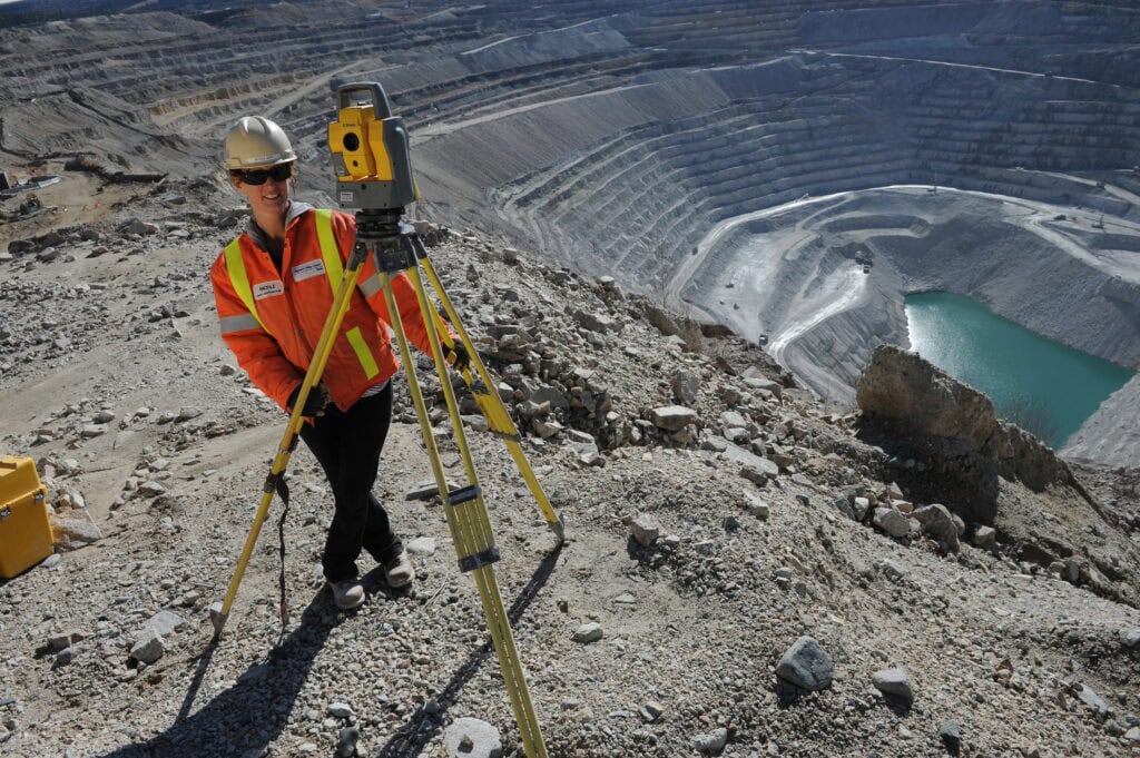 A person in a reflective vest is using survey equipment. Behind them is a deep valley with a surface copper mine and water at the bottom is visible.