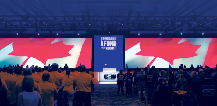In a large, darkened convention space, crowds of people are standing facing the front, with their backs to the camera. Lit up on the giant screens, is a close-up of a giant Canadian flag, waving. The same image is projected on both screens. There is a podium between the screens with a USW logo on it.