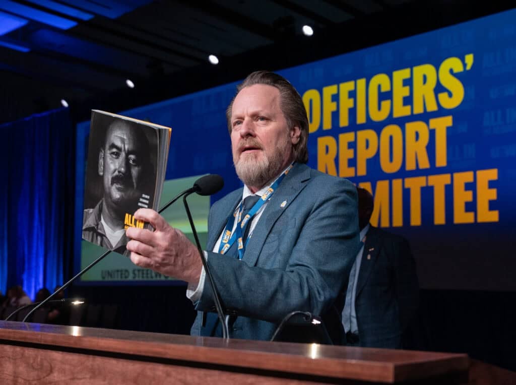 A bearded man is shown from the chest up in a suit and tie, speaking into a microphone at a large event with a giant screen behind him showing partial elements of the words Officers’ Report. He is holding up a magazine-style book with a glossy cover.