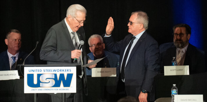 A man standing on a stage on the right with his right hand raised facing another man who is reading from a text.