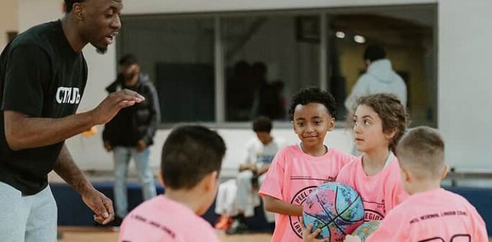 Four kids in pink t-shirts run a basketball drill with an adult coach in an indoor gym setting.