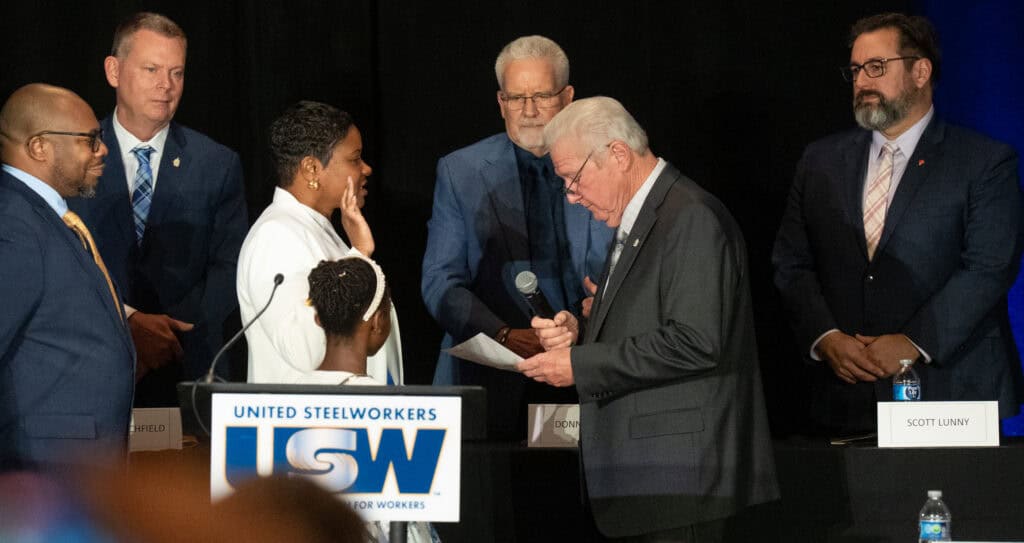 A woman and a young girl, seen from profile behind a podium, are facing a man holding a microphone, who is reading a document in front of him. The woman and the young girl are wearing white clothes, and the adult woman has her right hand raised. Another man observes the scene in the background.