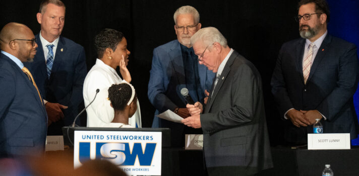 A woman and a young girl, seen from profile behind a podium, are facing a man holding a microphone, who is reading a document in front of him. The woman and the young girl are wearing white clothes, and the adult woman has her right hand raised. Another man observes the scene in the background.