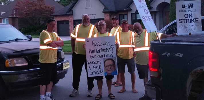 Six men wearing yellow shirts striped with high-visibility sashes stand between two trucks, holding posters that say "USW Local 14241 on strike" and "Passionate philantropist? But can't give workers a fair contract?" Next to the last message is the smiling headshot of a man wearing square glasses.