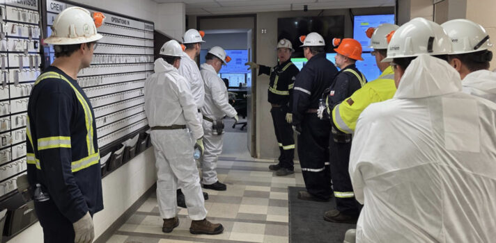 A group of ten workers in an interior space, facing away from the camera and towards a door to another room. They are in an industrial setting, wearing coveralls, some all navy and some all white, all wearing white hardhats with headlamps.