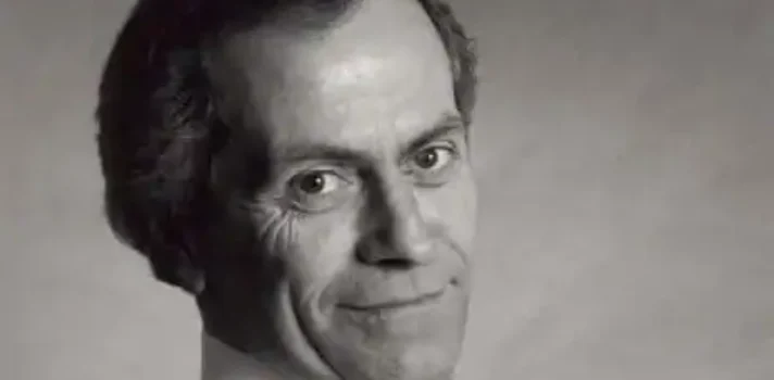 A black-and-white headshot of a man in a white shirt with a dark tie, taken from the side. He is leaning back slightly wtih his arms crossed.