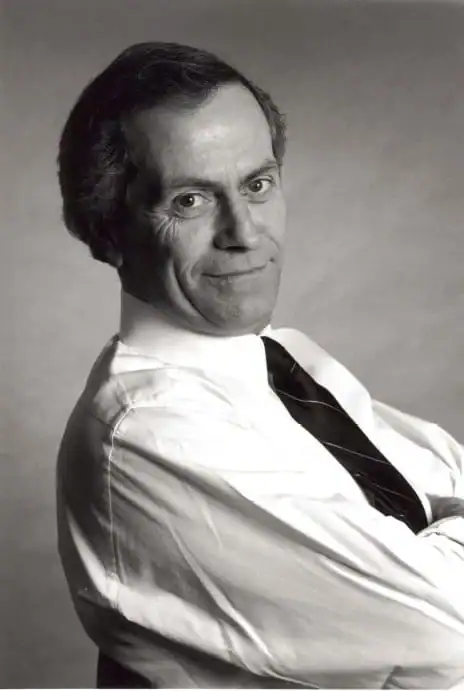 A black-and-white headshot of a man in a white shirt with a dark tie, taken from the side. He is leaning back slightly wtih his arms crossed.