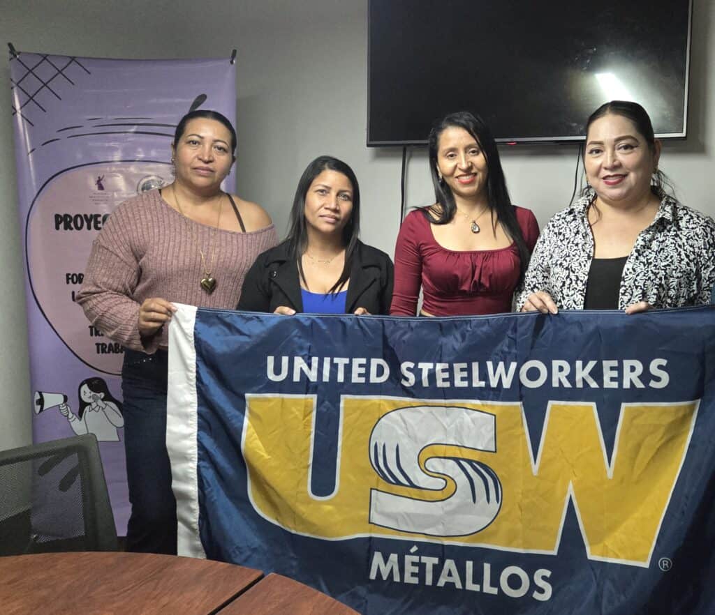 Four women stand together indoors holding a large United Steelworkers (USW) Métallos flag. A project banner is partially visible in the background.