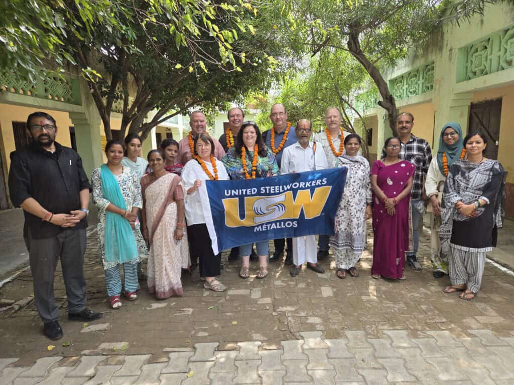A group of people pose outdoors in a courtyard, with several wearing orange flower garlands. People at the centre hold a United Steelworkers (USW) Métallos flag.