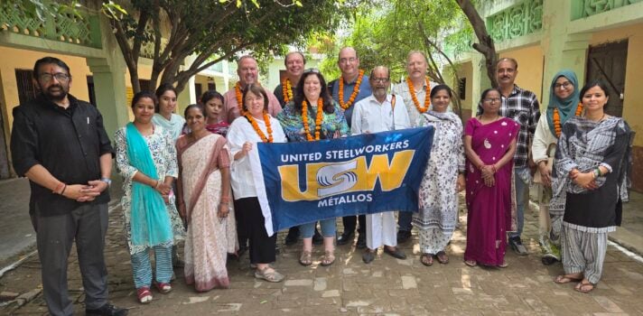 A group of people pose outdoors in a courtyard, with several wearing orange flower garlands. People at the centre hold a United Steelworkers (USW) Métallos flag.